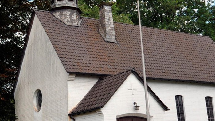 Weiße Kirche mit hohem Turm und braunem Dach, umgeben von Bäumen und Büschen