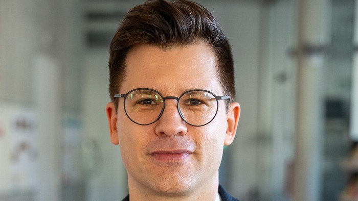 Ein Mann in einem dunklen Blazer und mit Brille steht mit einem professionellen, freundlichen Gesichtsausdruck in einem Innenraum.