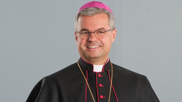 Ein Geistlicher in schwarzer kirchlicher Amtstracht mit einer rosa Bischofsmütze und Brille, der lächelt.
