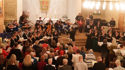 Et fællesskabskor og orkester optræder sammen i en stor, lysoplyst kirkehal under en aftenkoncert.