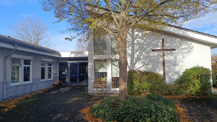 Moderne Kirchengebäude mit einem Kreuz an der Außenwand, umgeben von gestalteter Grünanlage und einem Baum.