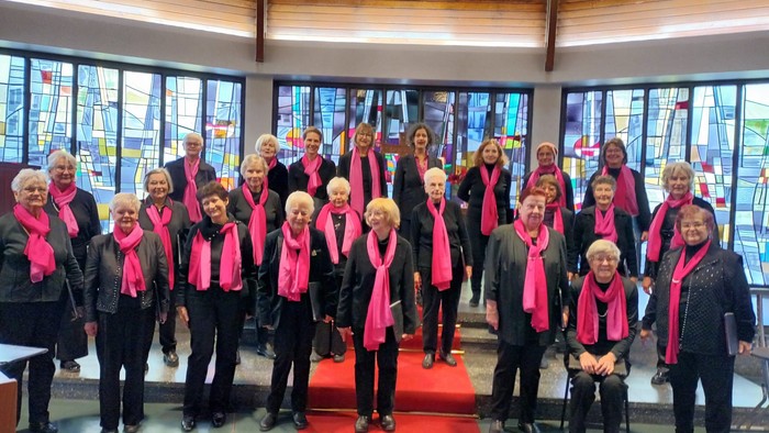 Eine Gruppe älterer Personen mit rosa Schals steht drinnen für ein Gruppenfoto zusammen.