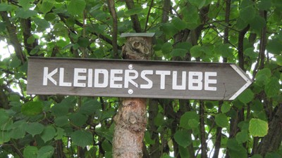 Holzwegweiser mit der Aufschrift „Kleiderstube“, umgeben von üppigem grünem Blattwerk.
