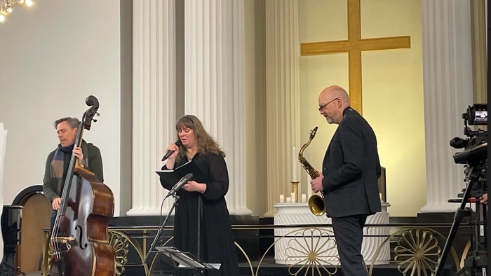 En kvinde synger i et kirkeinteriør, mens to mænd spiller bas og saksofon.