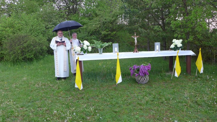Zwei Personen in weißen Anzügen stehen unter einem Regenschirm vor einem Tisch mit Blumen im Freien.