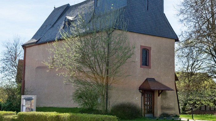 Kirche mit hohem Turm und Kreuz, umgeben von Bäumen und Gras.