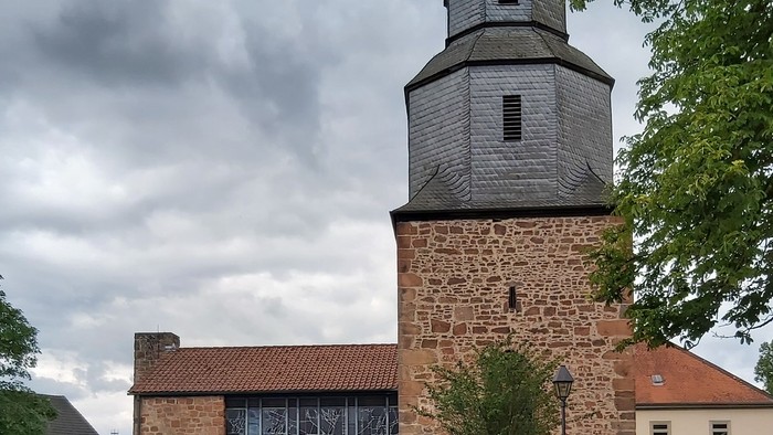Historische Steinkirche mit modernem Glasanbau, umgeben von Grünflächen und vor bewölktem Himmel gelegen.