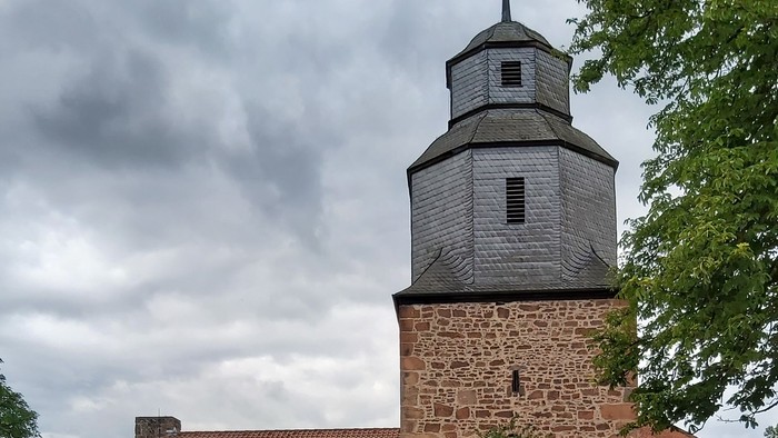 Historische Steinkirche mit modernem Glasanbau, umgeben von Grünflächen und vor bewölktem Himmel gelegen.