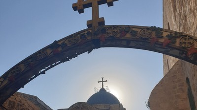 Ein steinernes Torbogen mit einem Kreuz rahmt eine historische Kirchenkuppel ein, die in goldenes Sonnenlicht getaucht ist.