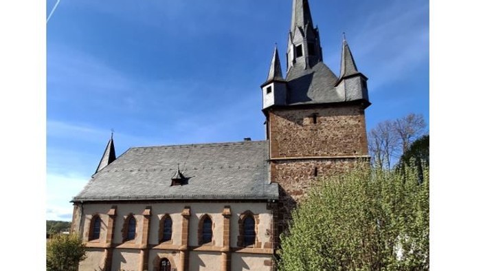 Kirche mit hohem Turmspitz und Rundbogenfenstern, umgeben von grünem Gras und Sträuchern.