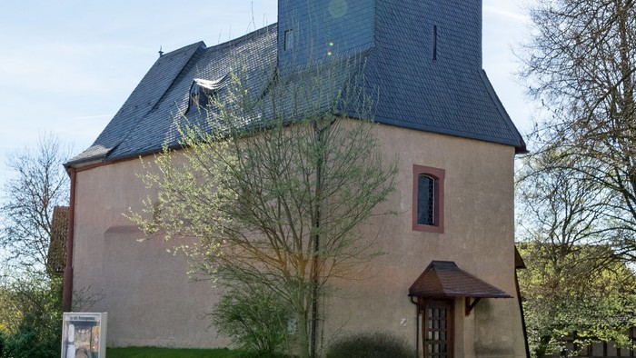 Kirche mit hohem Turm und Kreuz, umgeben von Bäumen und Gras.