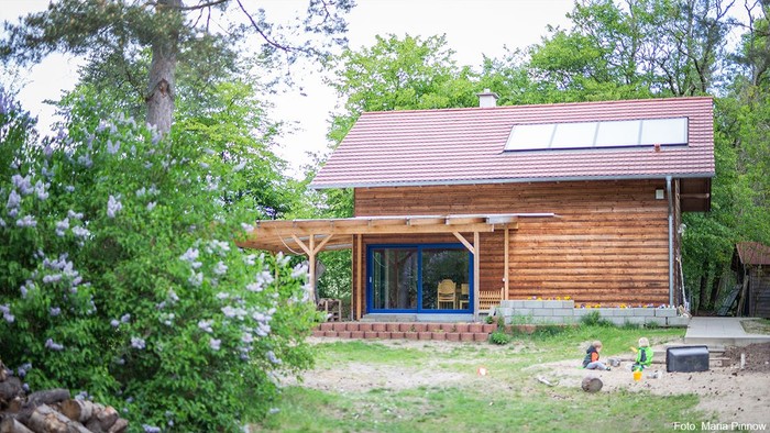 Holzhaus, davor ein Sandkasten mit spielenden Kindern und Sträucher.