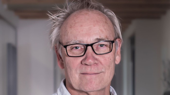 En mand med briller og gråt hår iført en lys knapskjorte indendørs.