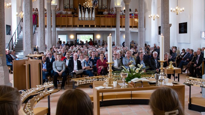 En stor kirkefuld med mennesker ved gudstjeneste, med orgel og lysstake.