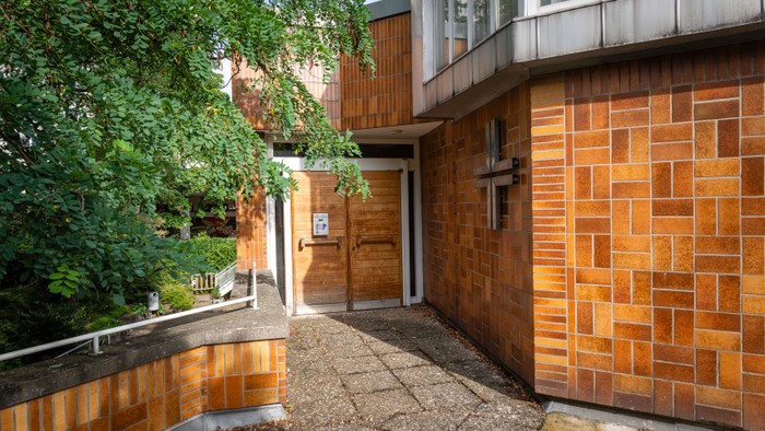 Das Bild zeigt einen Gehweg neben einem Gebäude mit Ziegelmauerwerk und Bäumen.