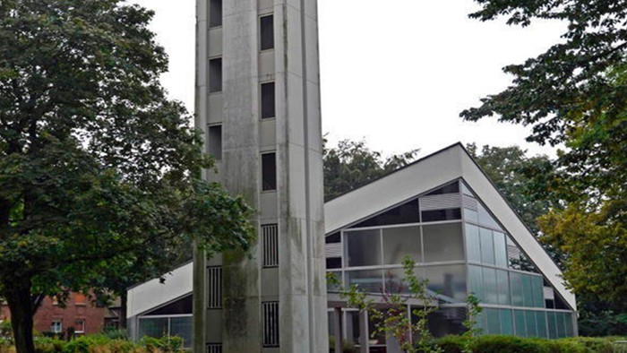 Das Bild zeigt eine moderne Kirche mit hohem Turm und Glasfront, umgeben von Bäumen.