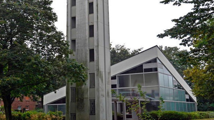 Das Bild zeigt eine moderne Kirche mit hohem Turm und Glasfront, umgeben von Bäumen.