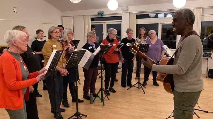 En gruppe mennesker, der synger sammen i et kor, ledet af en dirigent med noder.