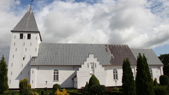 Nørre Løgum kirke, set fra syd med blå himmel med hvide skyer som baggrund. Kirken er hvidkalket, har et tårn og der er bly på taget. Forrest ses grønnehække og buske på kirkegården