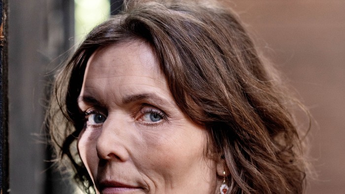 En kvinde med kort, bølget hår står tankefuldt ved et vindue med et neutralt udtryk.