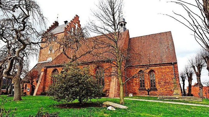 Alte Backsteinkirche mit hohem Turm und spitzem Dach, umgeben von Bäumen und Gras.