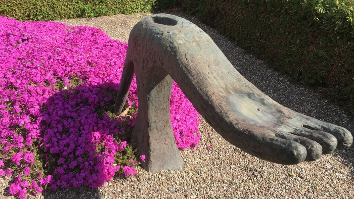 En stor bronzeskulptur af en fod, der stikker op af jorden mellem levende lyserøde blomster. Ved fostergraven