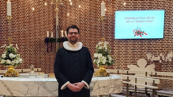 En mand i religiøst tøj står i en kirke med en bamse i hånden, omgivet af legetøj på gulvet.