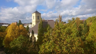 Eine friedvolle Kirche mit einem hohen Turm steht inmitten buntem Herbstlaub unter einem teilweise bewölkten Himmel.