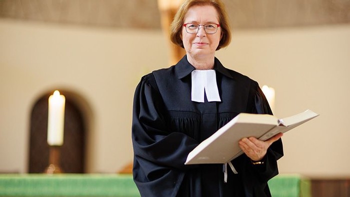 Eine Pfarrerin steht im Innenraum einer Kirche und hält ein aufgeschlagenes Buch in den Händen. Sie trägt eine schwarze Amtstracht mit Beffchen und blickt ruhig und aufmerksam in die Kamera. Im Hintergrund ist der Altarraum zu erkennen, mit Kerzenlicht u