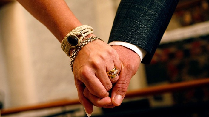 To personer, der holder hinanden i hånden, pyntet med ringe og armbånd, i en formel sammenhæng.