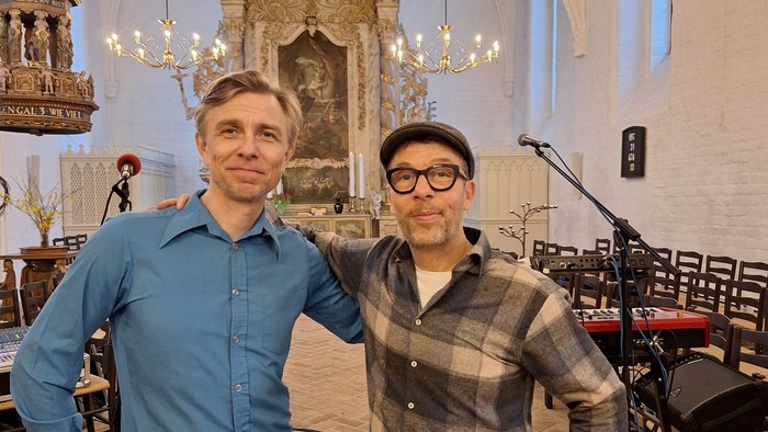 To mænd poser sammen i en kirke med musikudstyr og prangende dekorationer i baggrunden.