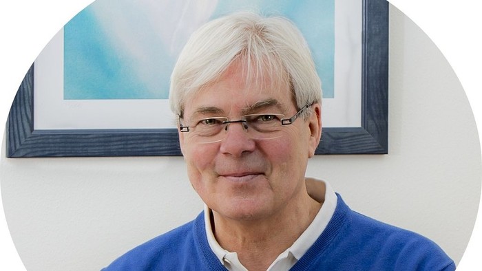 Mann mit weißen Haaren und Brille, der einen blauen Pullover trägt.