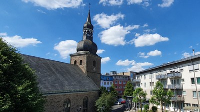 Historische Kirche mit hohem Kirchturm, umgeben von modernen Wohngebäuden unter einem strahlend blauen Himmel.
