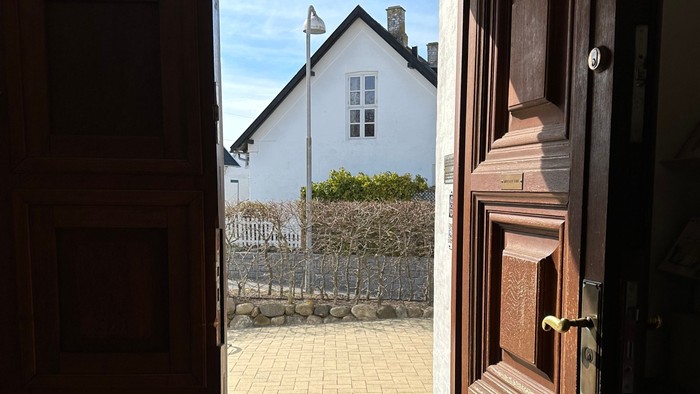 En åben dør viser ud til en stenlagt gård med et hvidt hus og blå vinduer.
