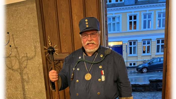 En mand i historisk militæruniform står og holder en ceremoniel stav nær indgangen til en gammel bygning.