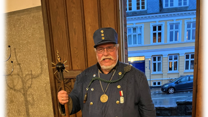 En mand i historisk militæruniform står og holder en ceremoniel stav nær indgangen til en gammel bygning.