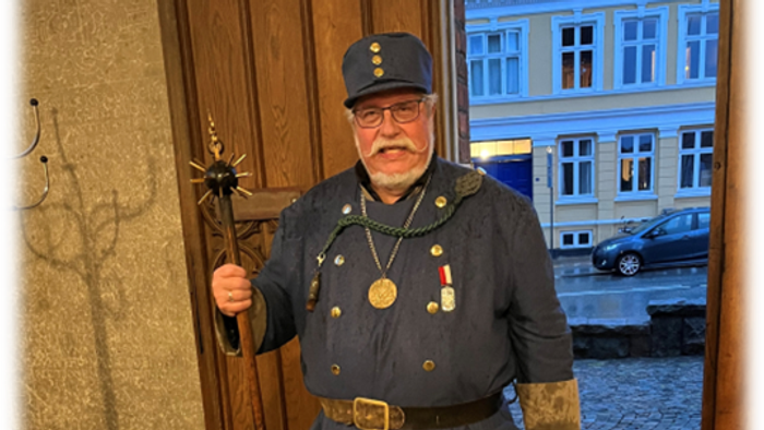 En mand i historisk militæruniform står og holder en ceremoniel stav nær indgangen til en gammel bygning.