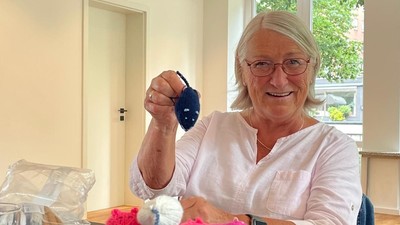 En glad kvinde holder en strikket mus, mens hun er omgivet af farverige strikkede legetøj på et bord.