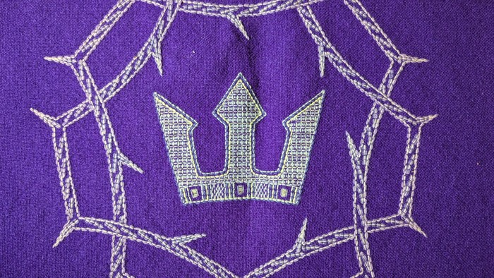 Komplexes weißes Kronen-Stickmuster auf violettem Stoff mit spinnwebenartiger Stickerei.