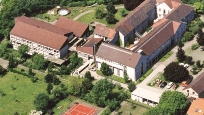 Ein großes Gebäude mit roten Dächern und einem Tennishirt im Vordergrund, umgeben von Grünflächen und Bäumen.