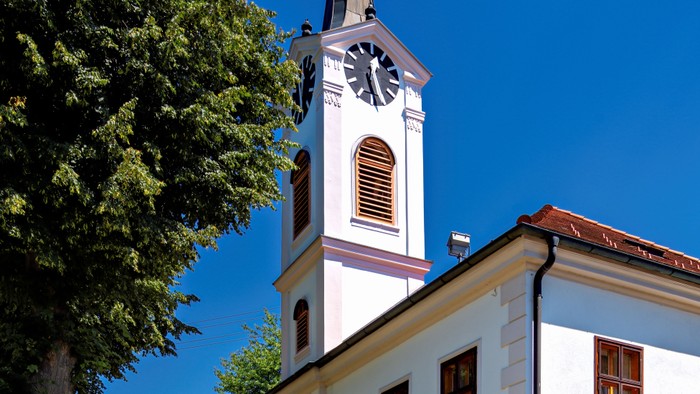 Weiße Kirche mit Uhrturm und rotem Dach, umgeben von Bäumen und Gras.
