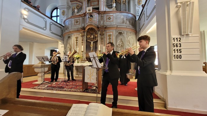 Eine Gruppe von Musiker:innen in formeller Kleidung tritt in einer prächtigen Kirche auf, spielt Blechblasinstrumente und singt.