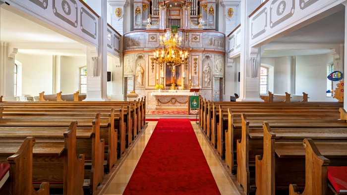 Kircheninnenraum mit hölzernen Kirchenbänken, rotem Teppich und Kronleuchter.