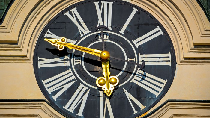 Elegante verzierte Uhr mit römischen Ziffern und goldenen Dekorzeigern vor einem cremefarbenen architektonischen Hintergrund.