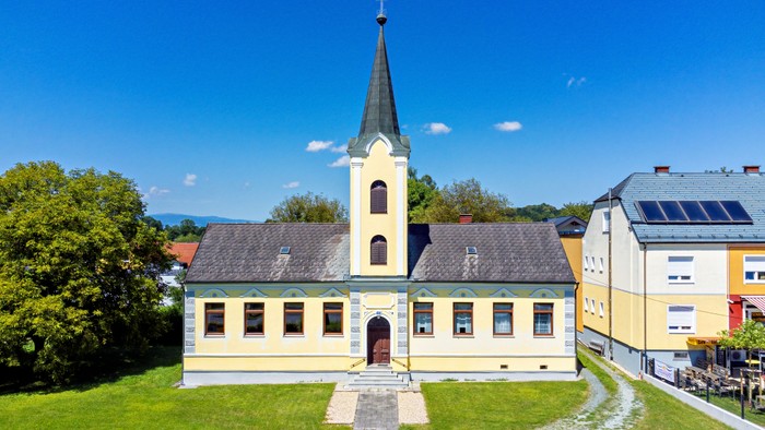 Kirche mit hohem Turm und gelbem Gebäude, umgeben von grünem Gras und blauem Himmel.