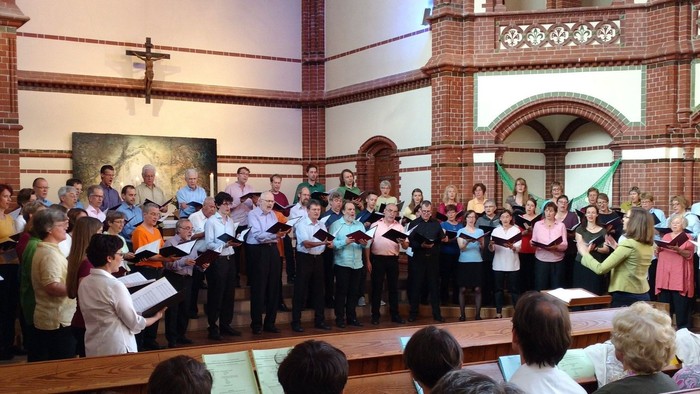 Ein großer Chor tritt in einer Kirche auf, dirigiert von einem Dirigenten, während Zuschauer im Hintergrund sitzen.