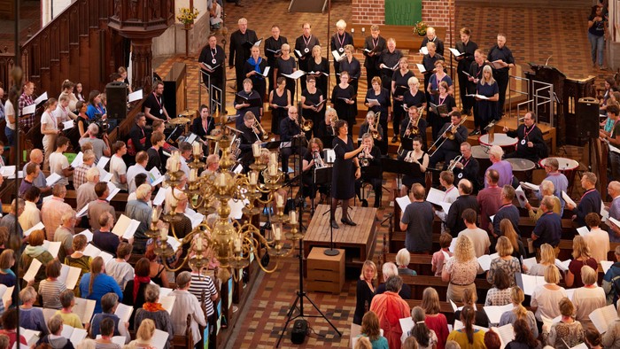 Ein großer Chor und ein Orchester treten gemeinsam in einer prächtigen, historischen Kirche auf, die mit aufmerksamen Zuhörern gefüllt ist.