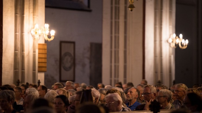 Eine überfüllte Indoor-Veranstaltung unter eleganten Kronleuchtern, bei der die Teilnehmer aufmerksam nach vorne blicken.