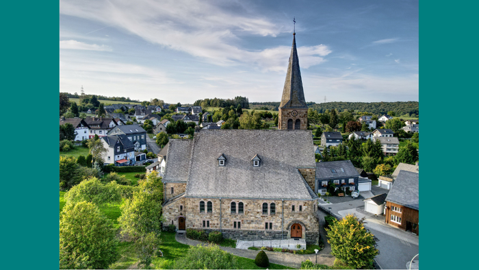 Ein Kirchengebäude mit hohem Turm in einem grünen, hügeligen Dorf.