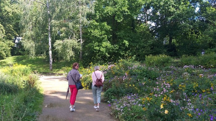 Zwei Personen schlendern einen üppigen, blumenbewachsenen Pfad entlang, umgeben von dichtem Grün und Bäumen.